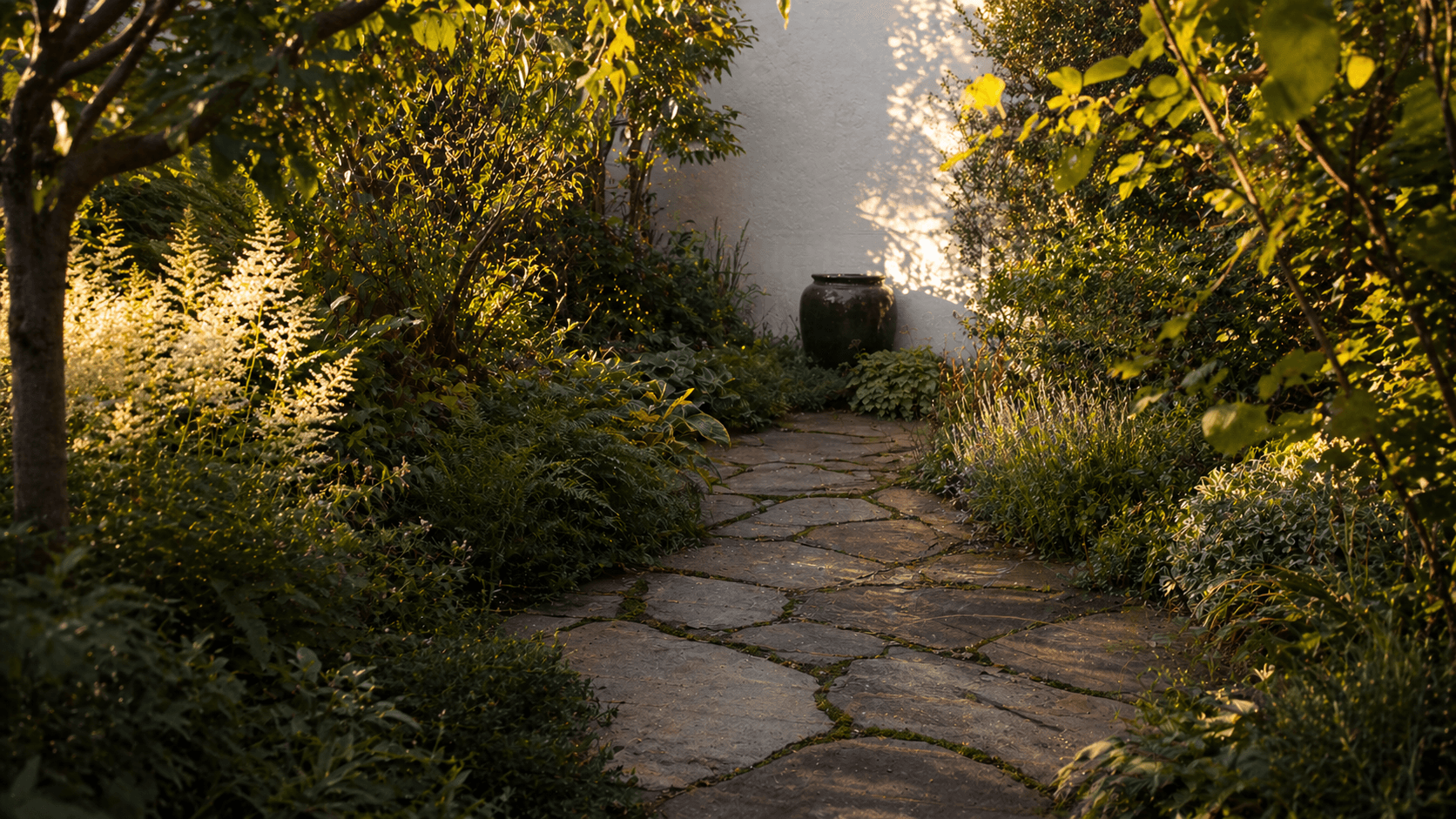 A beautifully layered small urban garden with diagonal paving, lush planting, and a focal point mirror on the far wall, photographed in warm evening light